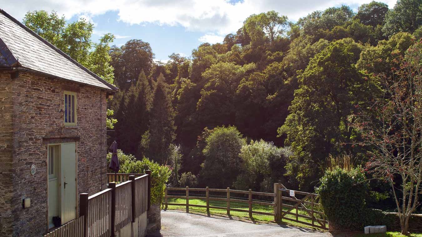 A rustic stone cottage with a slate roof sits beside a wooden fence, overlooking a lush green valley filled with dense trees under a partly cloudy sky.