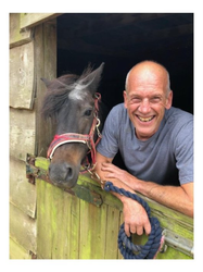 A smiling man leans on a wooden stable door beside a small dark pony wearing a red halter, both looking toward the camera.
