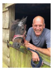A smiling man leans on a wooden stable door beside a small dark pony wearing a red halter, both looking toward the camera.