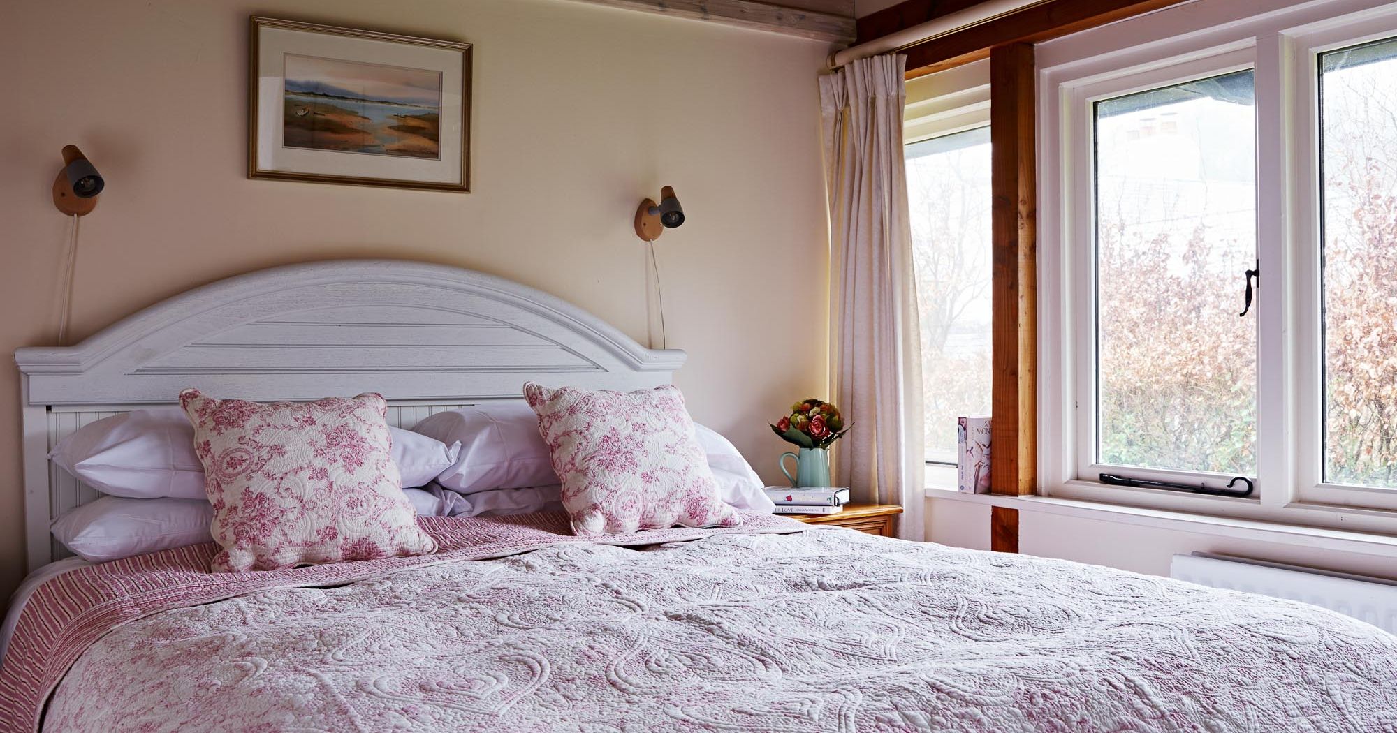 A cozy bedroom with a white wooden bed, pink floral bedding, and matching pillows. A large window with cream curtains lets in natural light, and a nightstand holds a book and flowers.