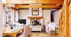 A bright, rustic living space with exposed wooden beams, a wood-burning stove, and striped sofas. A dining table set for breakfast sits near large windows with floral curtains.