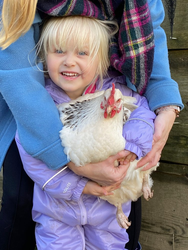 A smiling young girl in a purple snowsuit holds a white chicken with red wattles, with the support of an adult in a blue coat and plaid scarf standing behind her.
