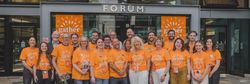 A group of people wearing matching orange 'gather' t-shirts standing and smiling in front of a building entrance labeled 'FORUM'.