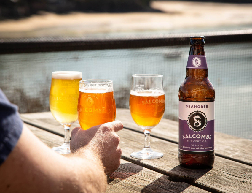A bottle of Salcombe Brewery Co. Seahorse amber ale on a wooden table outdoors, with three filled beer glasses in the background and a person's hand holding one glass.