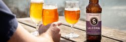 A bottle of Salcombe Brewery Co. Seahorse amber ale on a wooden table outdoors, with three filled beer glasses in the background and a person's hand holding one glass.
