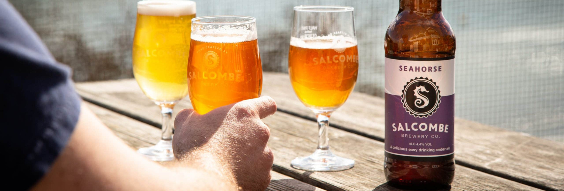 A bottle of Salcombe Brewery Co. Seahorse amber ale on a wooden table outdoors, with three filled beer glasses in the background and a person's hand holding one glass.