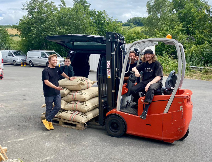 A group of four people smiling while operating a forklift to unload a stack of burlap sacks from a vehicle in an outdoor parking area, with green trees and a cloudy sky in the background.