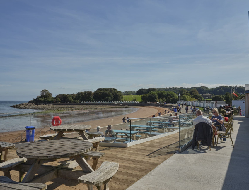 A seaside view with wooden picnic tables and outdoor seating on a deck overlooking a sandy beach, people walking along the shoreline, and green trees under a clear blue sky.