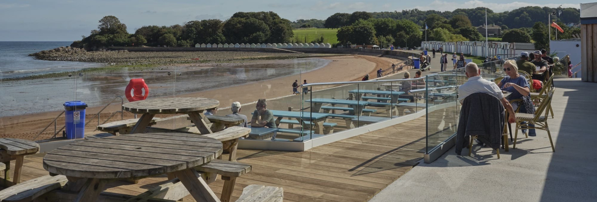 A seaside view with wooden picnic tables and outdoor seating on a deck overlooking a sandy beach, people walking along the shoreline, and green trees under a clear blue sky.