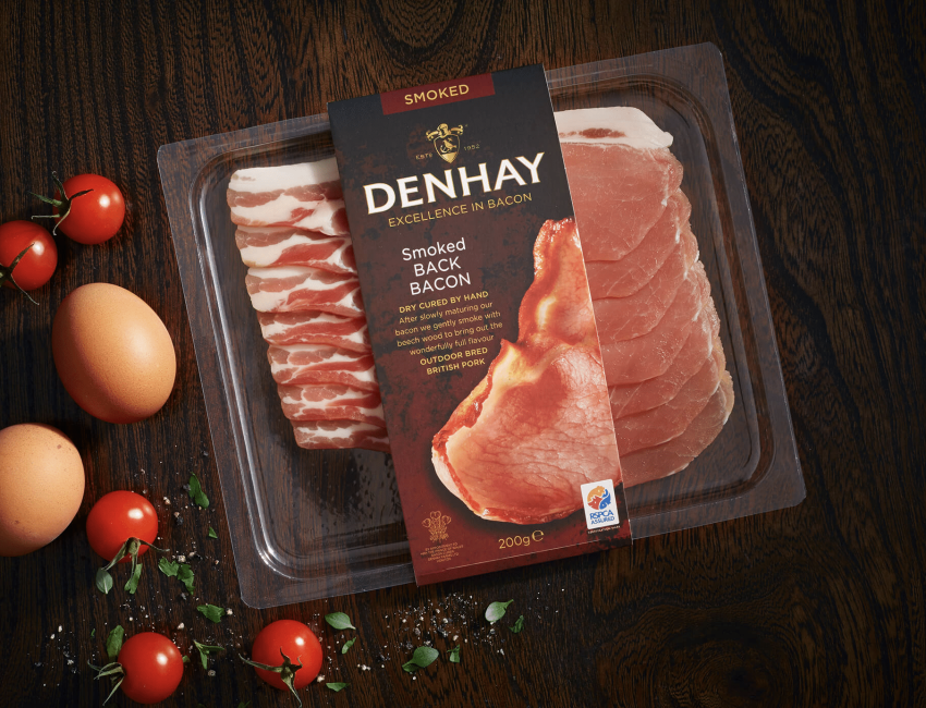 A pack of Denhay smoked back bacon placed on a dark wooden surface, surrounded by fresh cherry tomatoes and two brown eggs. The packaging highlights the bacon's quality and origin.