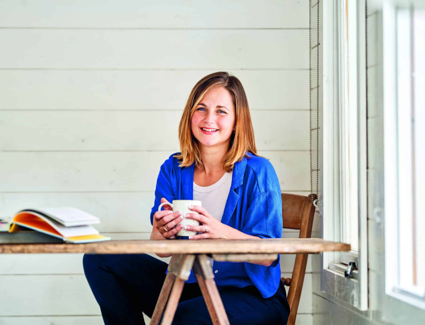 A woman in a blue shirt smiles while sitting at a wooden table with an open book and holding a mug. She is in a bright room with white wooden walls and a window to her right.
