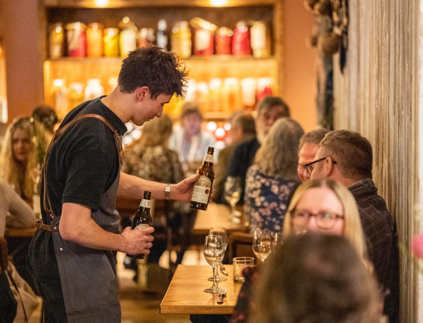 A waiter in a dark apron serves bottled drinks to a table of customers in a cozy, warmly lit restaurant. Other diners are visible in the background, enjoying their meals.