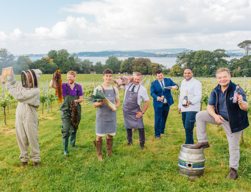 Six men and one woman in a vineyard display various local products, including honey, seaweed, leeks, meat, wine, and beer. The scenic background includes fields, trees, and a distant lake.