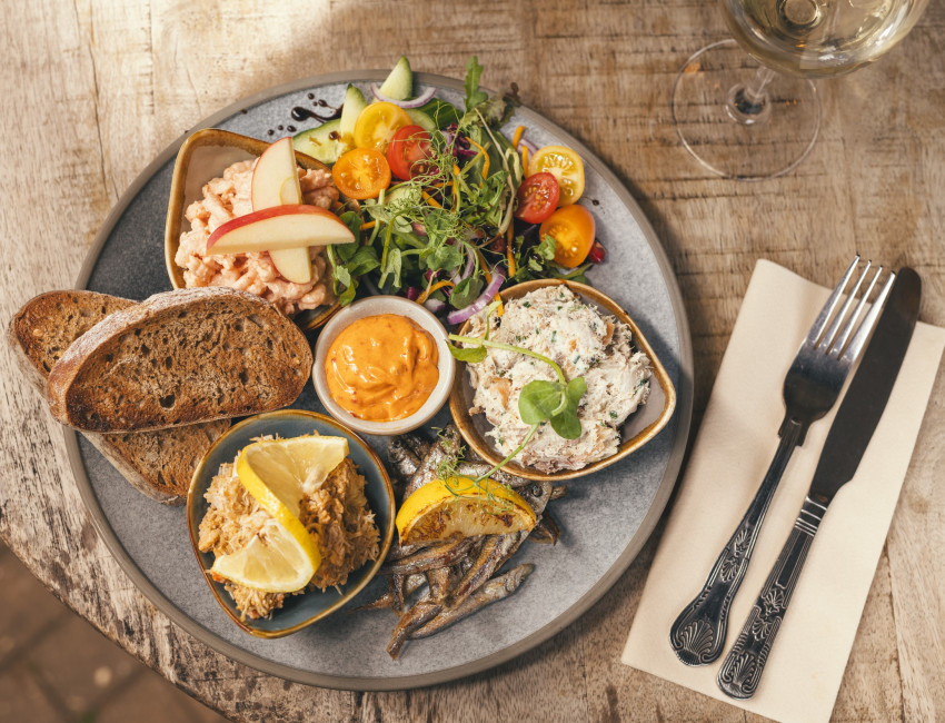 A seafood platter with various dishes, including shrimp with apple slices, smoked fish spread, crab with lemon, anchovies, cherry tomatoes salad, and toasted bread, served on a rustic table