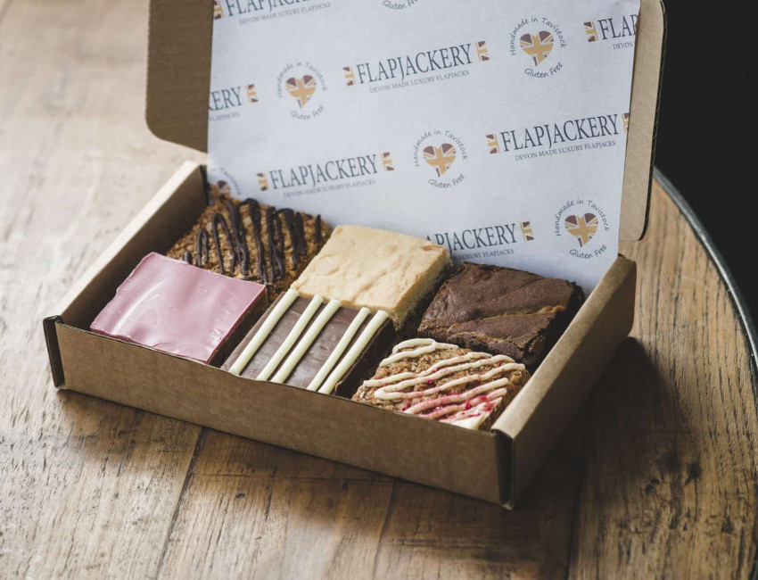 A cardboard box containing an assortment of six luxury flapjacks from Flapjackery, with various flavors and toppings, displayed on a rustic wooden table.