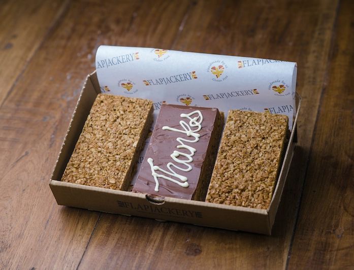 A box of three flapjacks from Flapjackery, featuring two plain oat bars and one chocolate-coated bar with 'Thanks' written in white icing, displayed on a wooden surface.