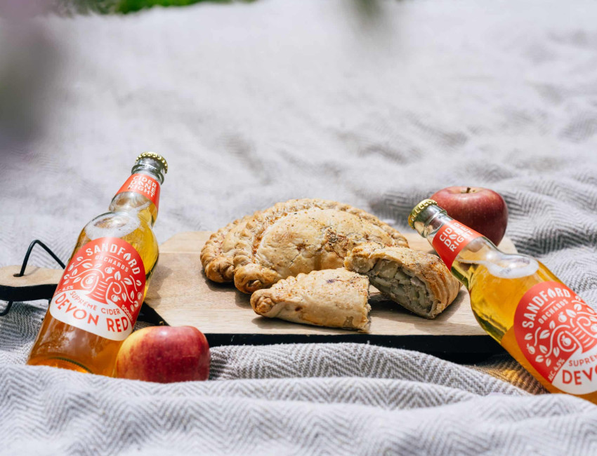 A picnic scene featuring two bottles of Sandford Orchards Devon Red cider, two apples, and three Cornish pasties, one sliced open, on a wooden board over a grey blanket.