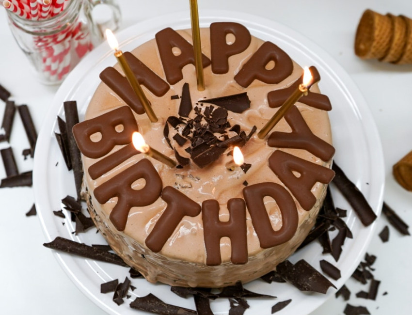 A chocolate ice cream cake with 'Happy Birthday' spelled out in chocolate letters, topped with lit candles and chocolate shavings, displayed on a white cake stand.
