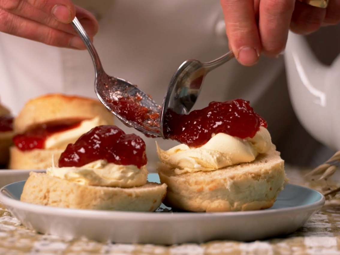 Hands spooning strawberry jam onto a cream-topped scone on a plate