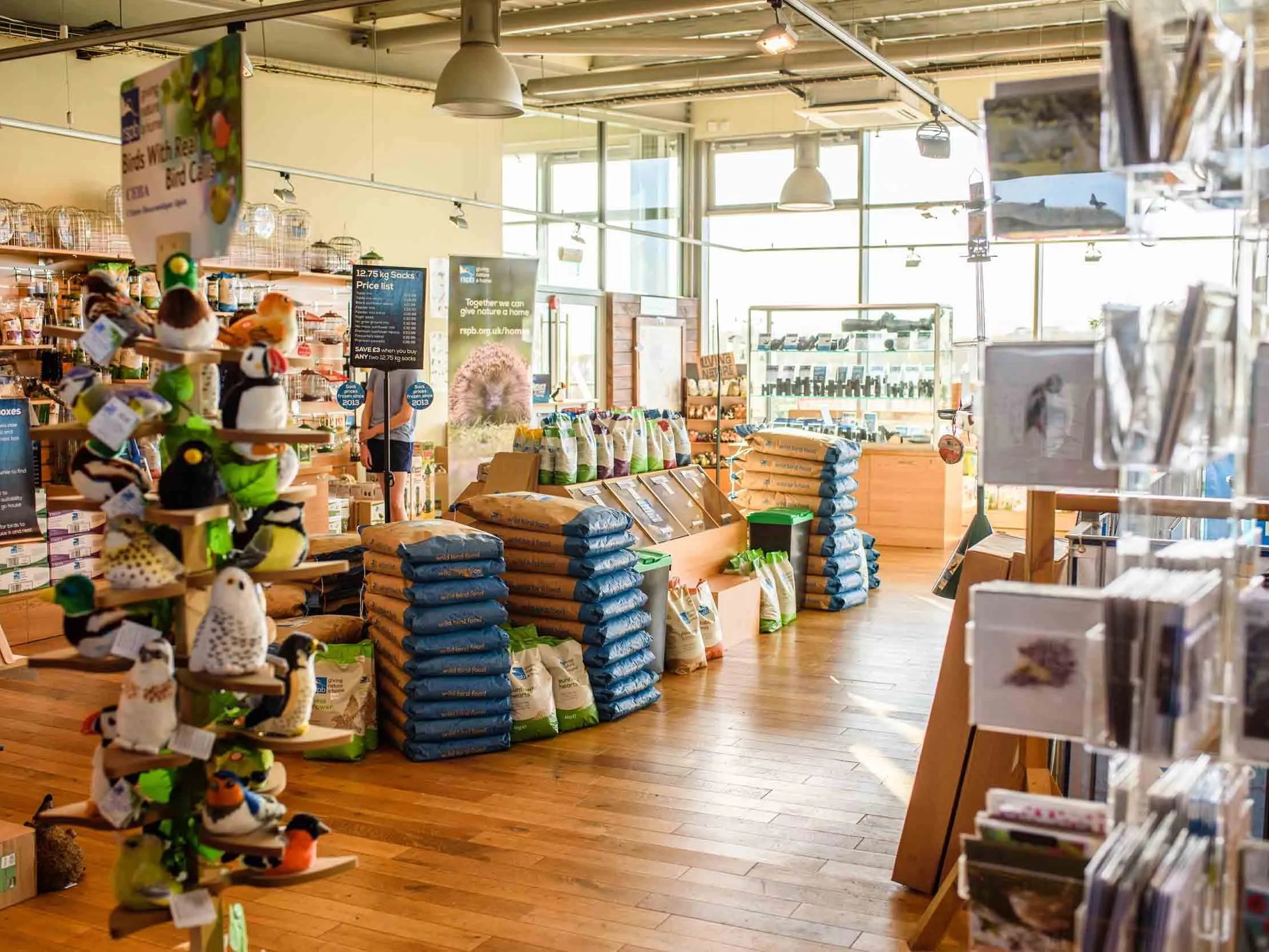 Bright shop interior with bird-themed gifts, stacks of bagged feed, and postcard racks by large windows
