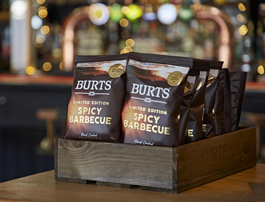 Several bags of Burts Limited Edition Spicy Barbecue hand-cooked potato chips displayed in a wooden box, with a blurred background featuring bokeh lights in a bar setting.
