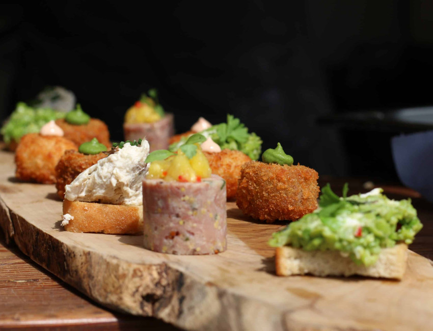 An assortment of gourmet appetizers on a rustic wooden board, including croquettes, topped with sauces, spreads, and garnishes, served on a wooden table.