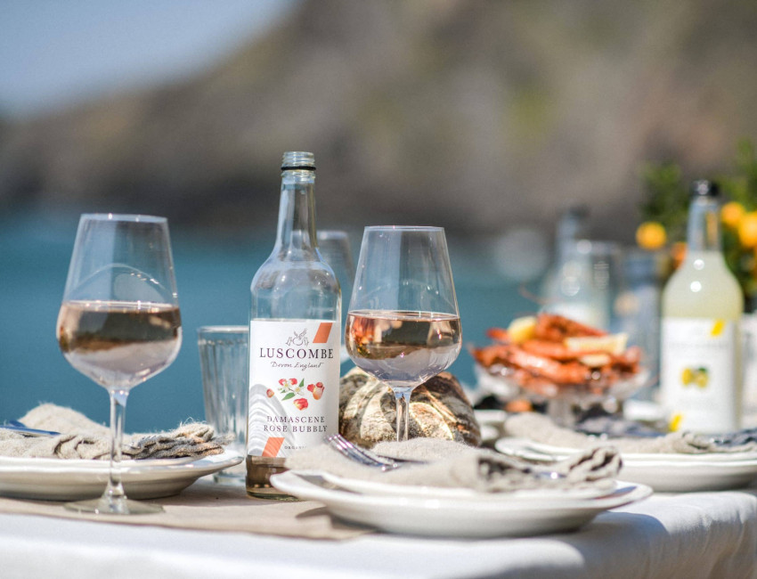 A seaside table setting with bottles of Luscombe drinks, glasses of rosé, seafood, and citrus decor, overlooking a blurred coastal background.