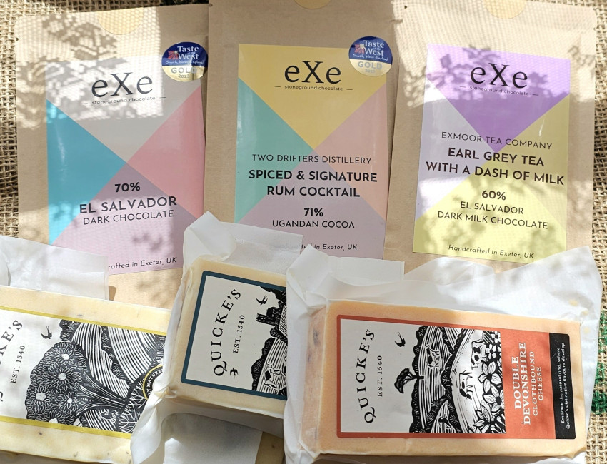 An assortment of artisan chocolate bars and cheese blocks by eXe and Quicke's, featuring various flavors like dark chocolate and Earl Grey tea on a burlap background.
