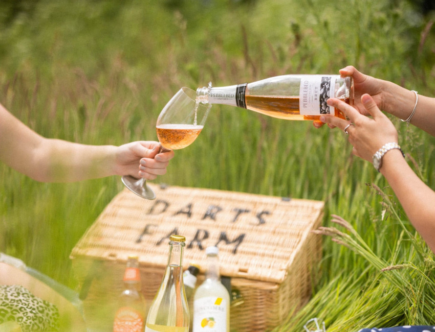 A picnic scene with two people in a grassy field, one pouring Pebbled Ridge rosé wine into a glass. A wicker basket labeled 'Darts Farm' and various drink bottles are visible.