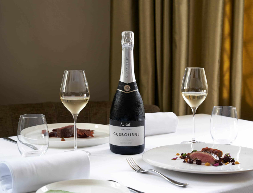 Elegant dining setup featuring a bottle of Gusbourne Blanc de Blancs wine with two filled glasses, served alongside two gourmet plated dishes on a white tablecloth.