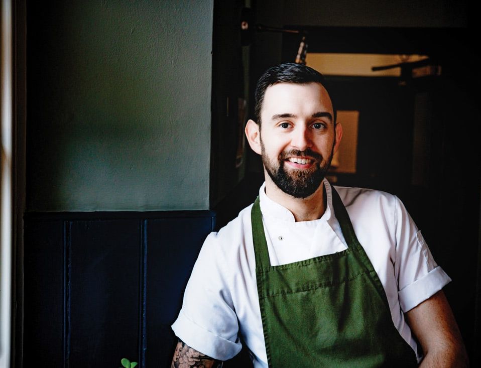 Portrait of a smiling chef with a beard wearing a white chef’s coat and a green apron, standing by a window in a warmly lit restaurant interior.