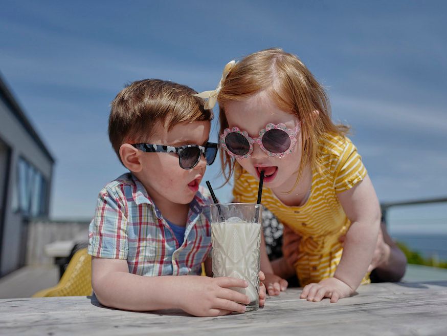 Two young children in sunglasses sharing a milkshake through straws at an outdoor table on a sunny day