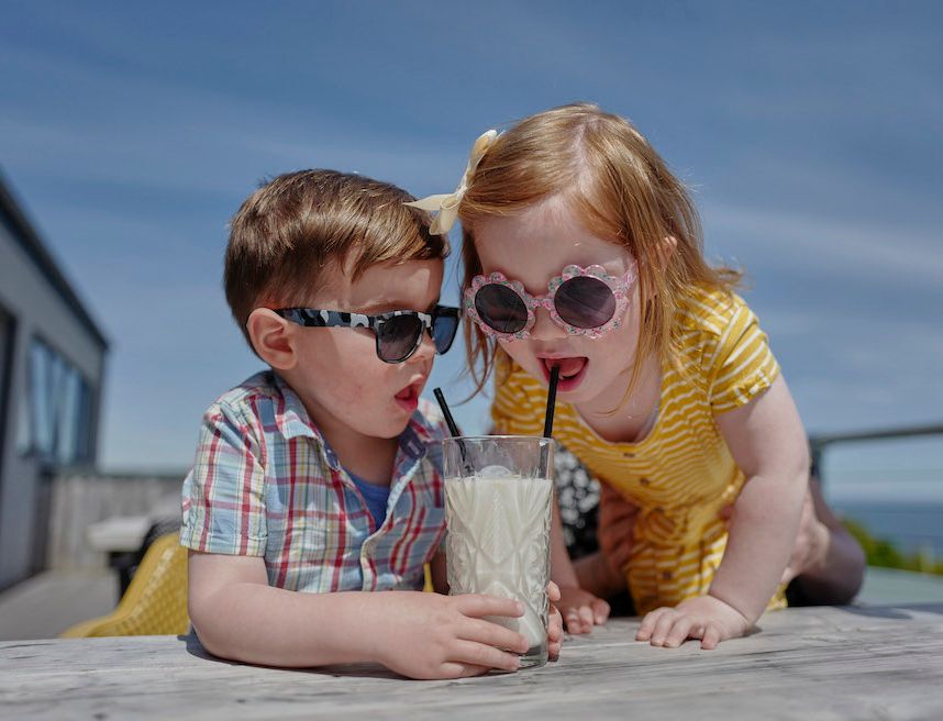 Two young children in sunglasses sharing a milkshake through straws at an outdoor table on a sunny day