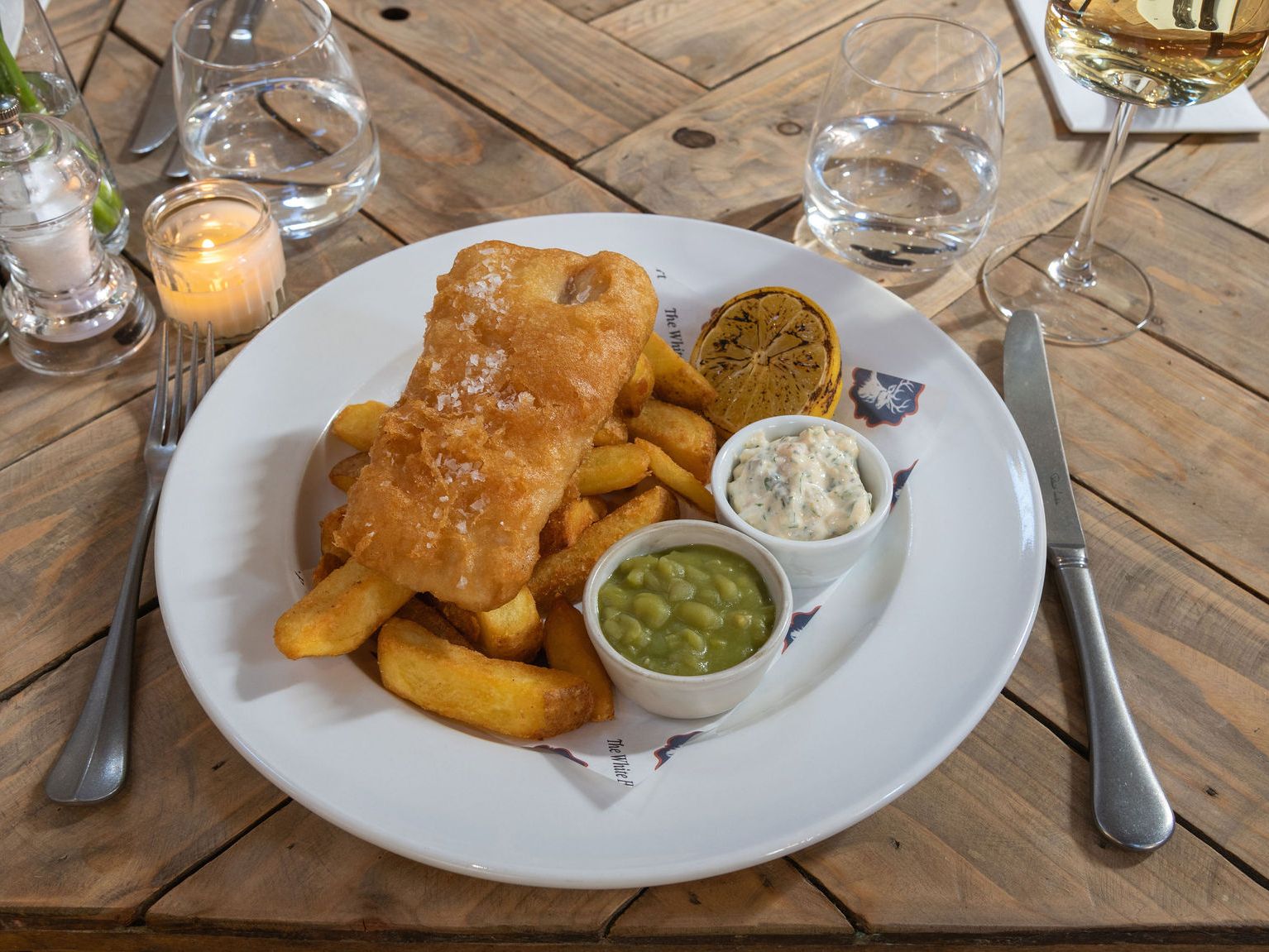 Plate of fish and chips with mushy peas, tartare sauce and a charred lemon wedge on a wooden table