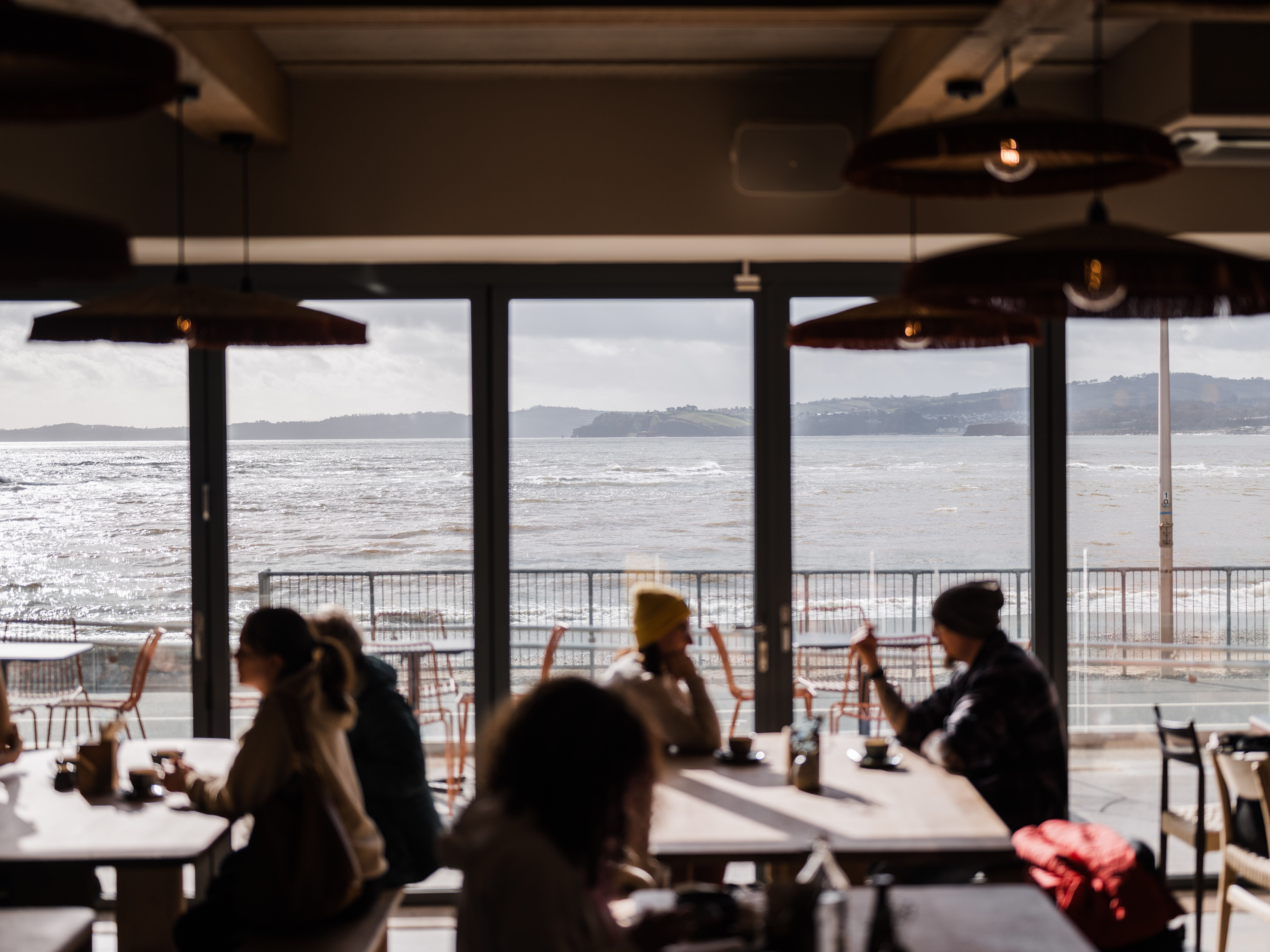 People sitting at tables in a café with large windows overlooking a bright, choppy sea and distant hills