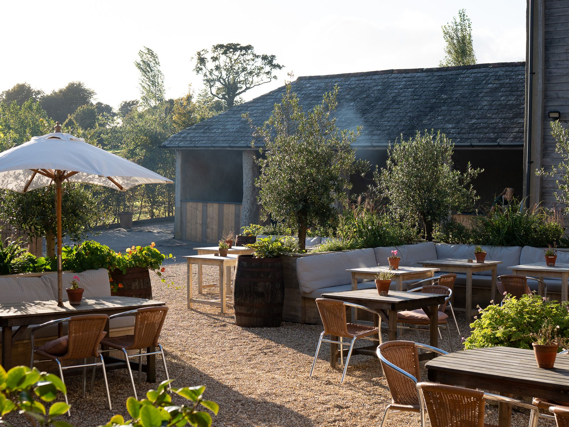 Outdoor seating area at Shilstone Fire Shed with wooden tables, wicker chairs, a white parasol and potted plants beside a rustic shed