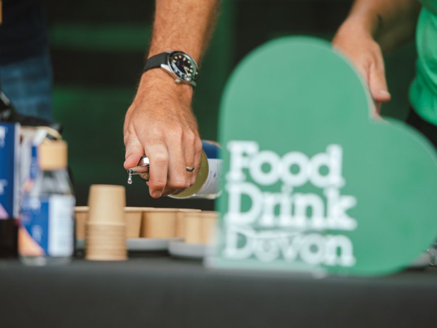 Greendale Love Local Food Festival image of hands pouring a cocktail sample next to a green Food Drink Devon heart logo
