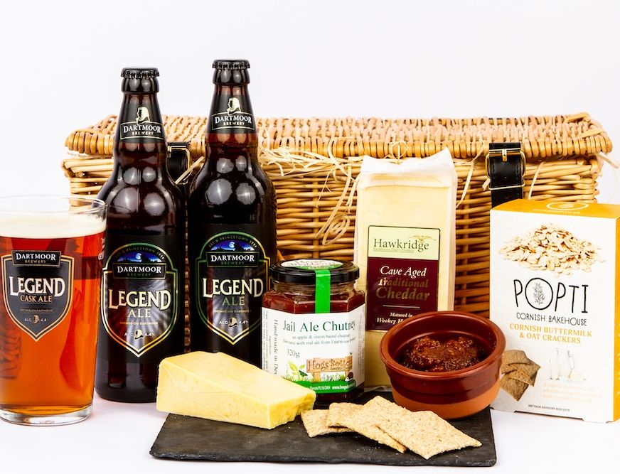 A hamper with two bottles and a pint of Dartmoor Legend Ale, cheddar cheese, chutney, oat crackers, and a small dish of chutney, set against a wicker basket background.