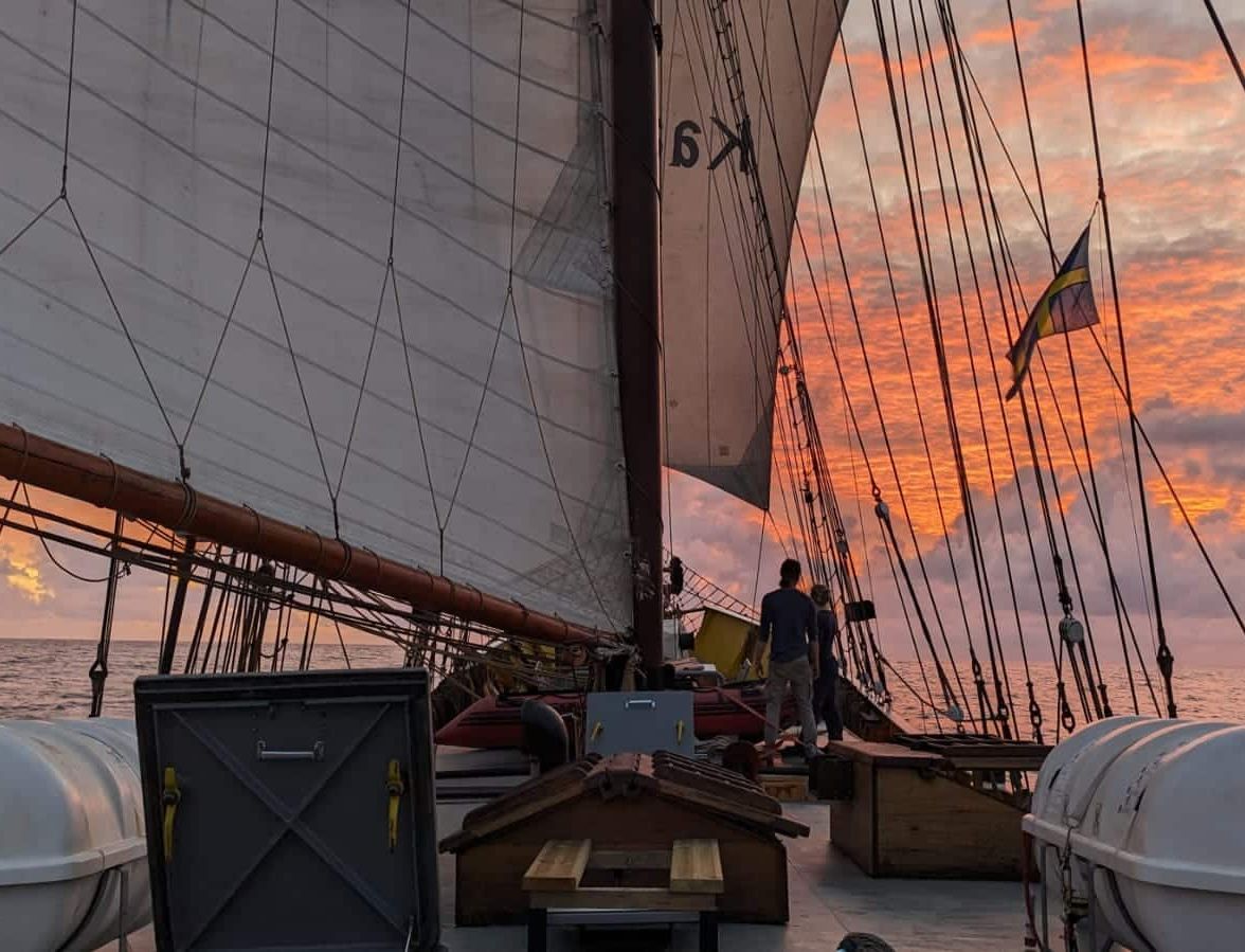 View from the deck of a sailing ship at sunset, with large sails overhead and a person standing near the stern, gazing at the orange and pink sky over calm ocean waters.