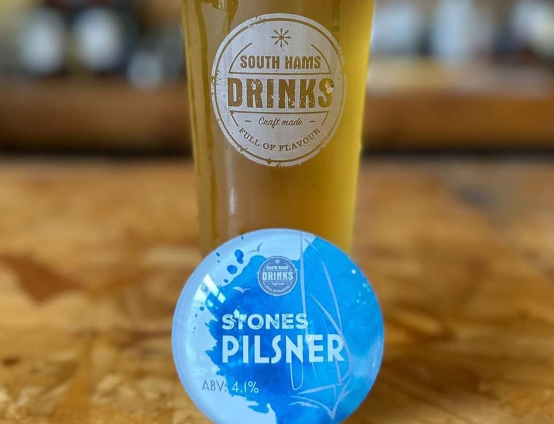 A pint of South Hams Drinks Stones Pilsner on a wooden table, with a logo on the glass and a blue circular label displaying 'Stones Pilsner, ABV: 4.1%' placed in front.