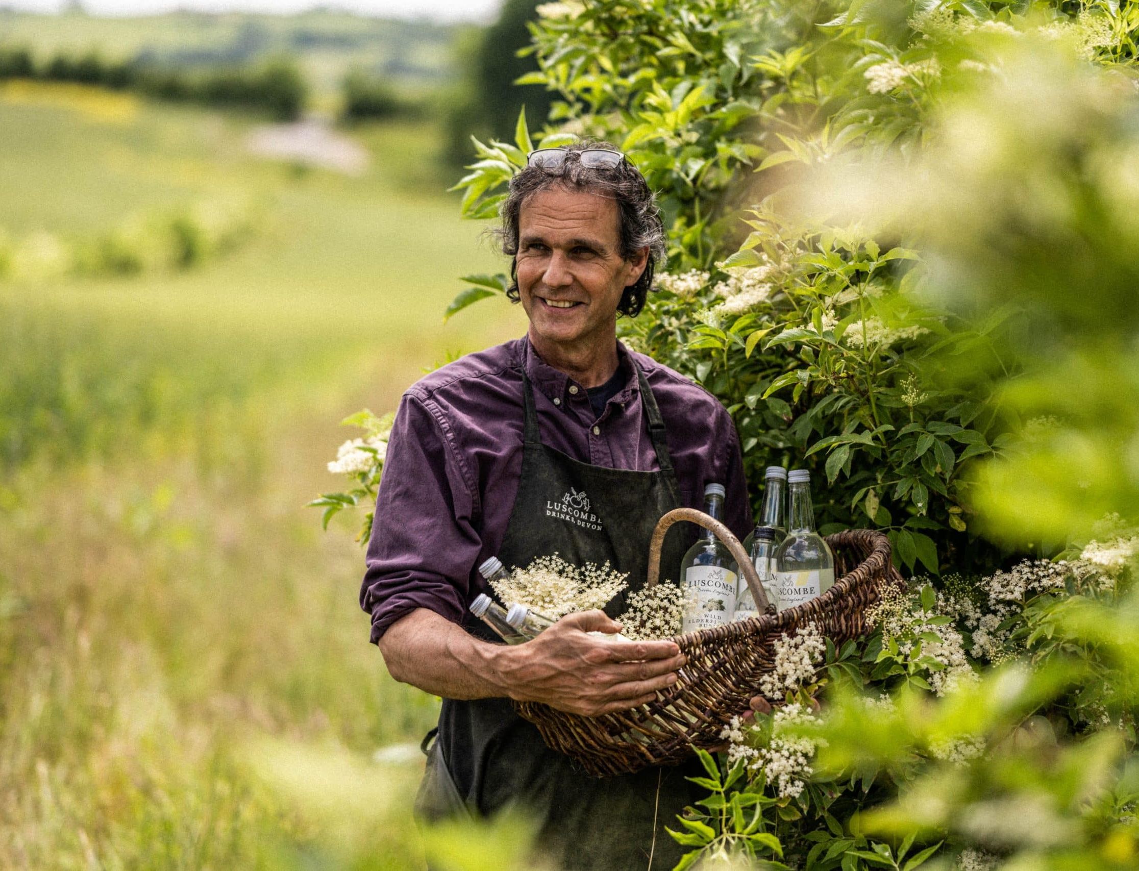 A man wearing a Luscombe apron stands in a field, holding a wicker basket filled with elderflowers and bottles of Luscombe drinks. Lush green foliage surrounds him under a bright sky.