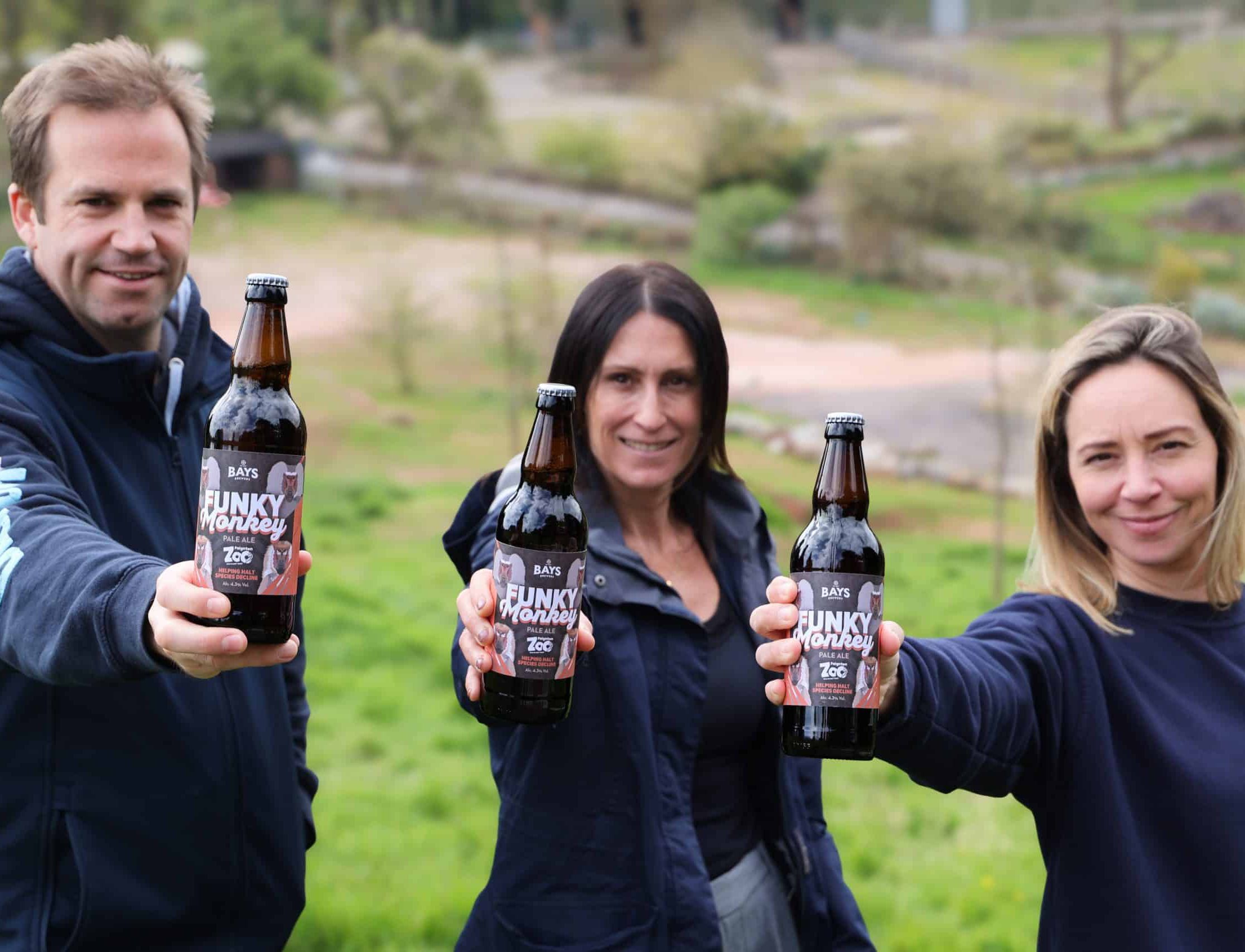 Three people smiling and holding bottles of Bays Funky Monkey Pale Ale, brewed in collaboration with Paignton Zoo, while standing outdoors in a grassy, scenic area.