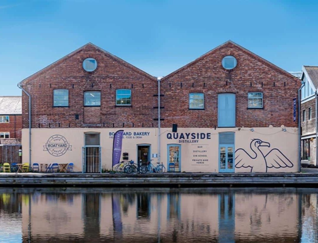 A view of two connected brick buildings by the water, housing Boatyard Bakery and Quayside Distillery, with outdoor seating, bikes, and a large bird mural on the distillery's facade.