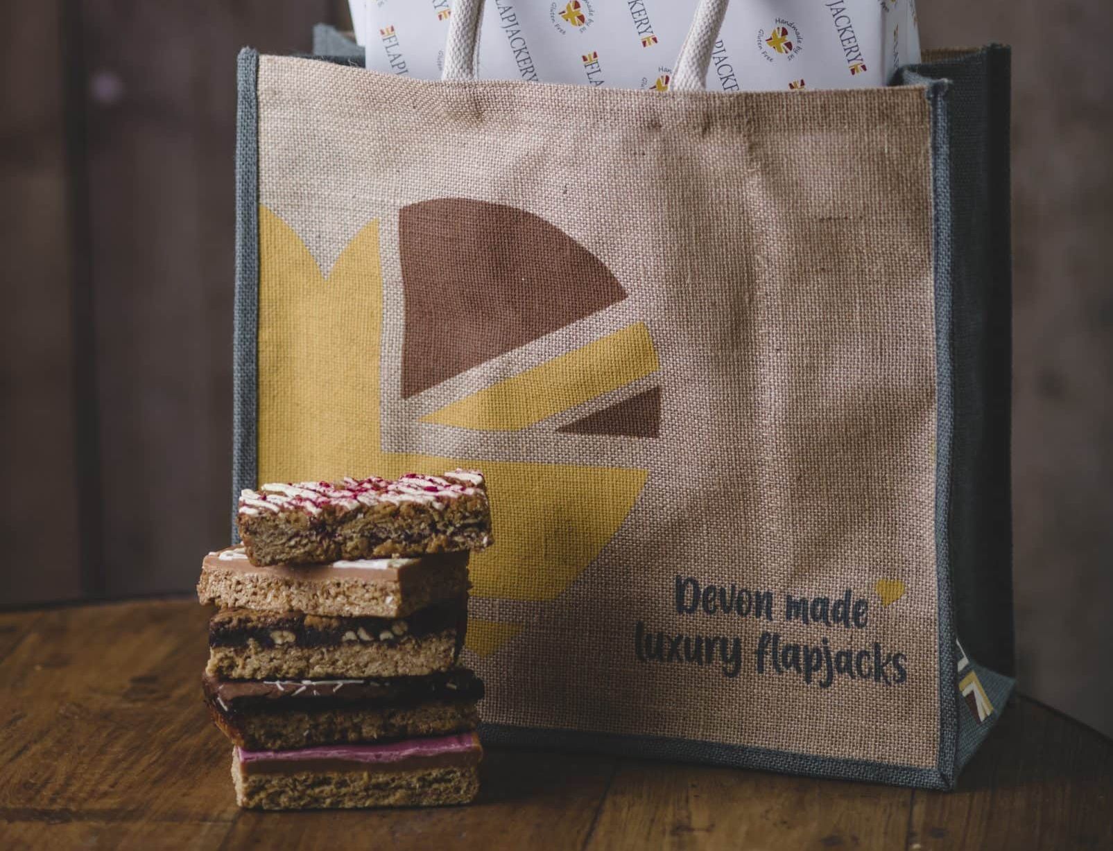 A jute bag labeled 'Devon made luxury flapjacks' filled with wrapped flapjacks and a gift voucher. A stack of assorted flapjacks is displayed in front of the bag on a wooden surface.