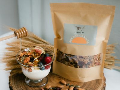 a westloaf granola pouch next to a bowl of granola, fruit and yoghurt being drizzled with honey
