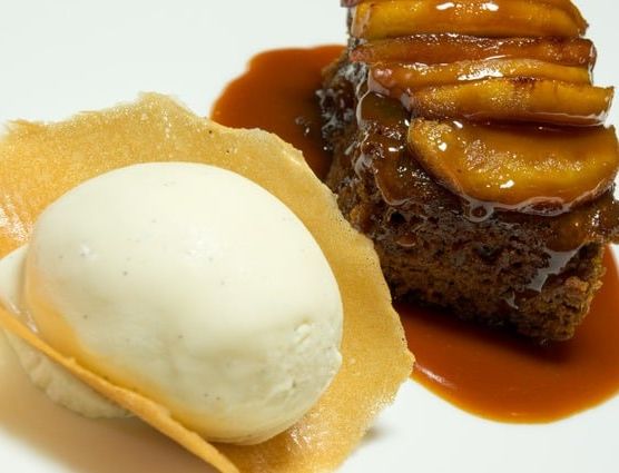 A dessert plate featuring a slice of caramel-glazed cake topped with caramelized apple slices is served with a scoop of vanilla ice cream resting on a thin, crispy wafer.