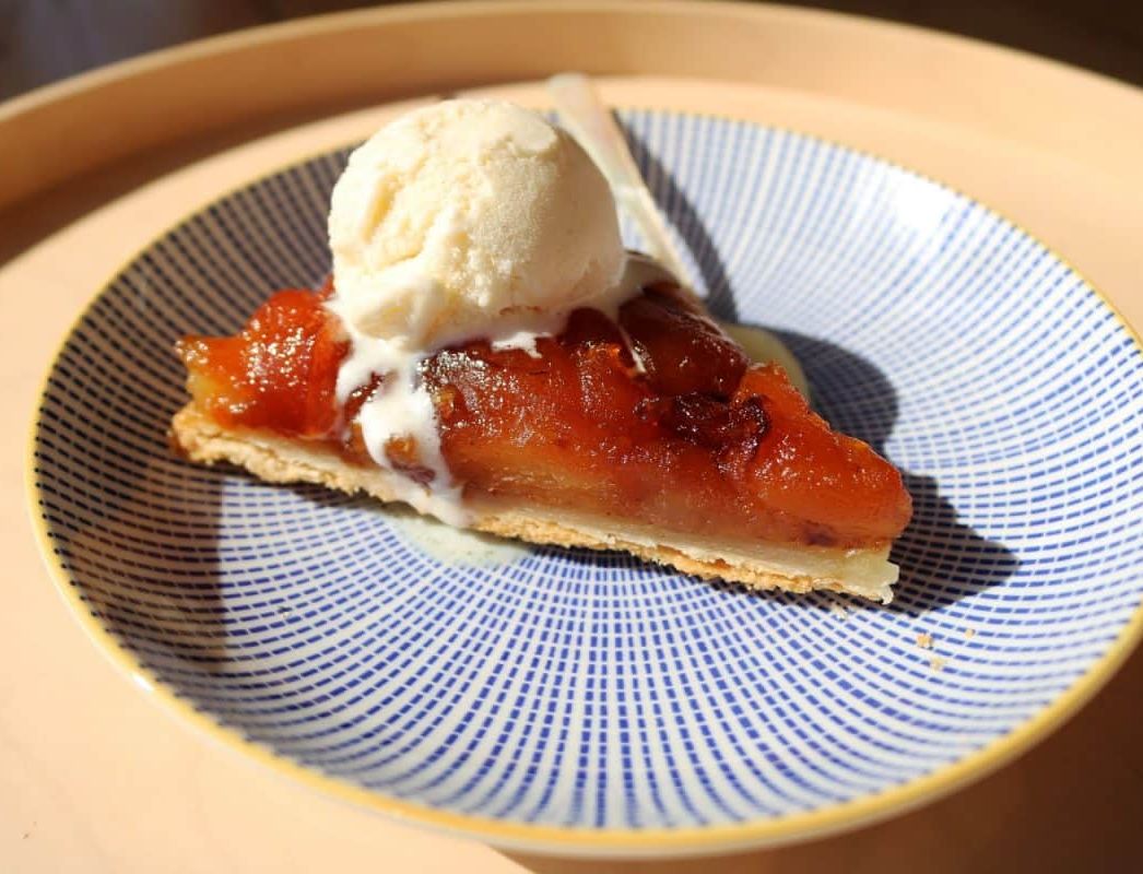 A slice of tarte Tatin, featuring caramelized apples on a flaky crust, is topped with a scoop of vanilla ice cream. The dessert is served on a blue patterned plate with a spoon.