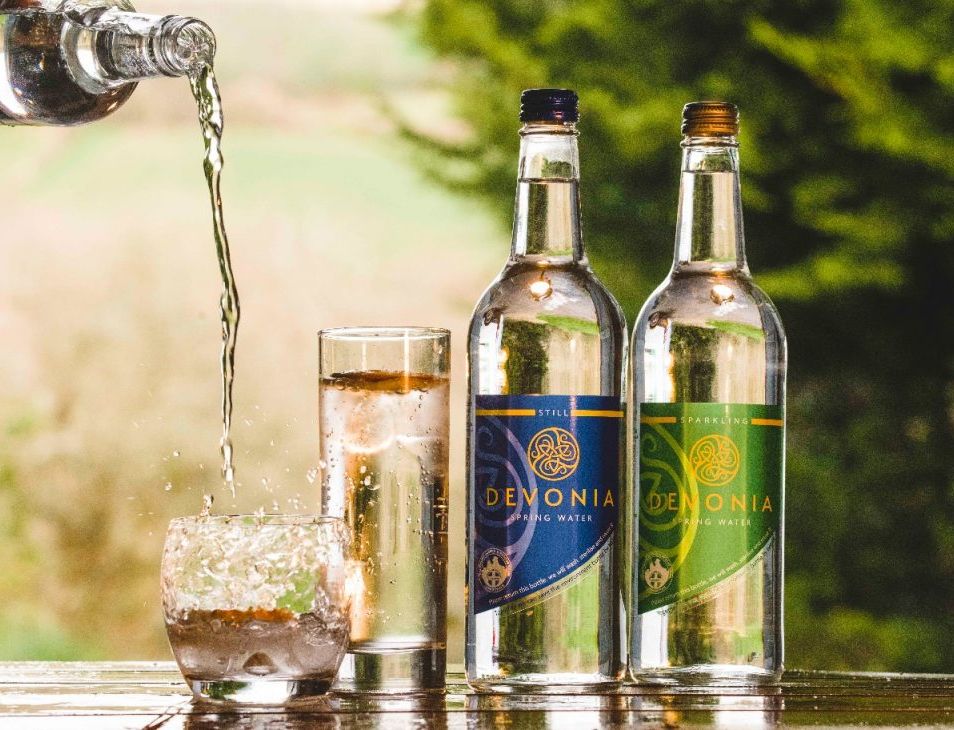 Water is being poured into a glass next to two bottles of Devonia spring water, one still and one sparkling. The scene is set outdoors with a blurred, green natural background.