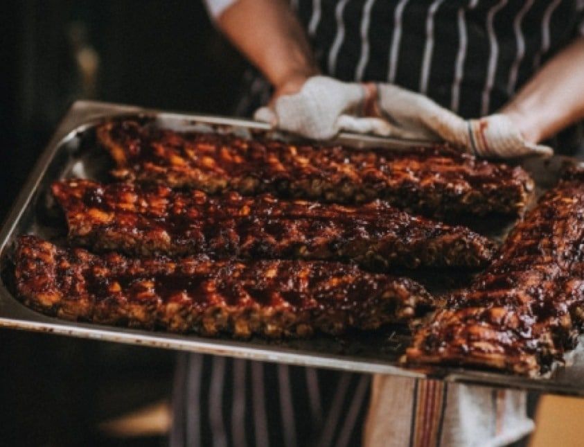 A person wearing an apron holds a tray of freshly cooked, glazed barbecue ribs. The ribs are richly coated in sauce, giving them a glossy appearance, ready to be served.