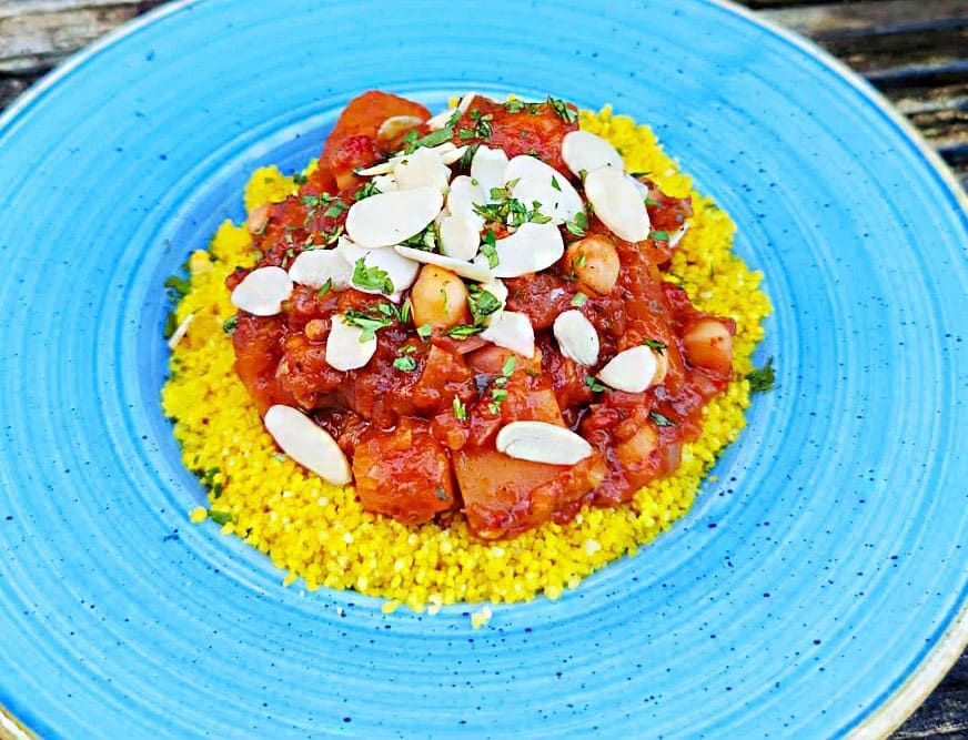 A vibrant plate of yellow couscous topped with a rich tomato-based vegetable stew, garnished with sliced almonds and fresh herbs, served on a bright blue dish.
